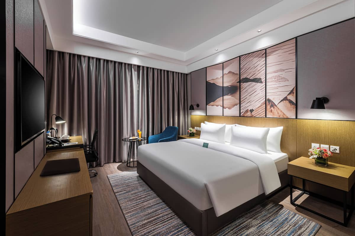 A modern hotel room with a large bed, a desk, a chair, a TV, and a wall with artwork.