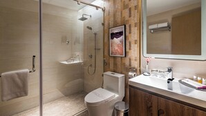 Un baño moderno con ducha envuelta en vidrio, inodoro, lavamanos con artículos de aseo y un cuadro en la pared.