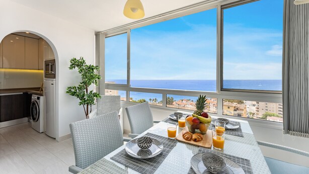 A modern kitchen with a washing machine, a dining area with a table set for breakfast, and a large window overlooking the sea. - Coastal Charm: Renovated 2-Bed Apt, Pool, Sea Views REF056 (Benalmádena)