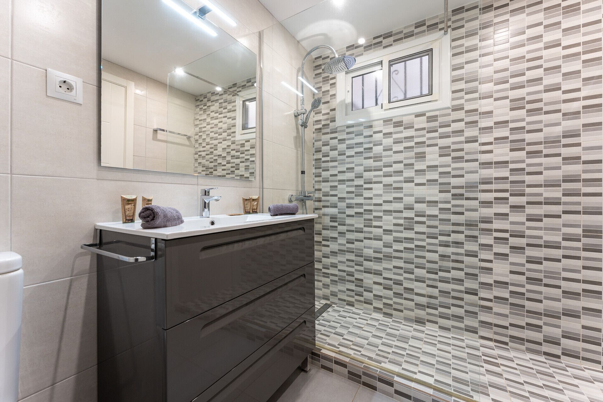 A modern bathroom with a large mirror, two sinks, a shower, and a tiled wall with a geometric pattern.
