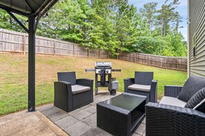 An outdoor patio with wicker furniture, a grill, and a view of a backyard with a wooden fence and trees. - Fire Pit, Central to RTJ, Downtown Auburn & Opelika - Sleeps 12 (Opelika)