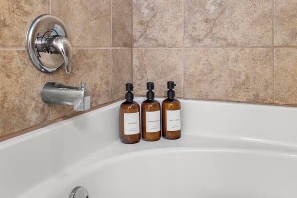 A bathroom with a bathtub, a faucet, and three bottles of shampoo, conditioner, and body wash. - Fire Pit, Central to RTJ, Downtown Auburn & Opelika - Sleeps 12 (Opelika)