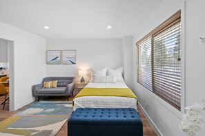 Ein Schlafzimmer mit einem Bett, einem grauen Sofa, einem blauen Hocker, einem Nachttisch und einem Fenster mit Jalousien.