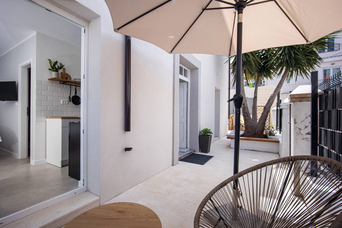 A modern kitchen with white tiles, a black refrigerator, and a wooden countertop. There is a large umbrella providing shade over a wicker chair and table.