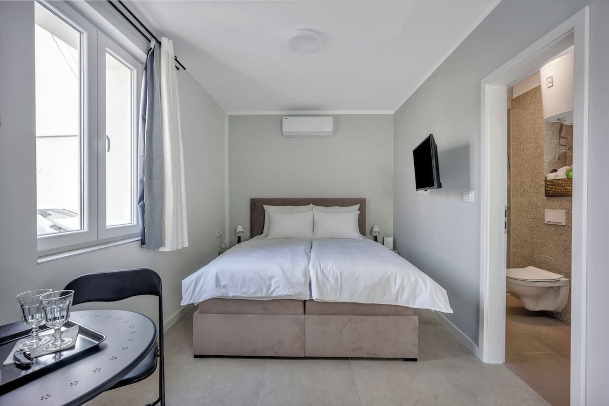 A bedroom with a bed, a window, a table with glasses, and a bathroom visible through an open door.