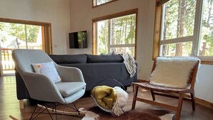 Ein Wohnzimmer mit grauem Schaukelstuhl, einer schwarzen Couch, einem Holztisch und einem Kuhfellteppich.