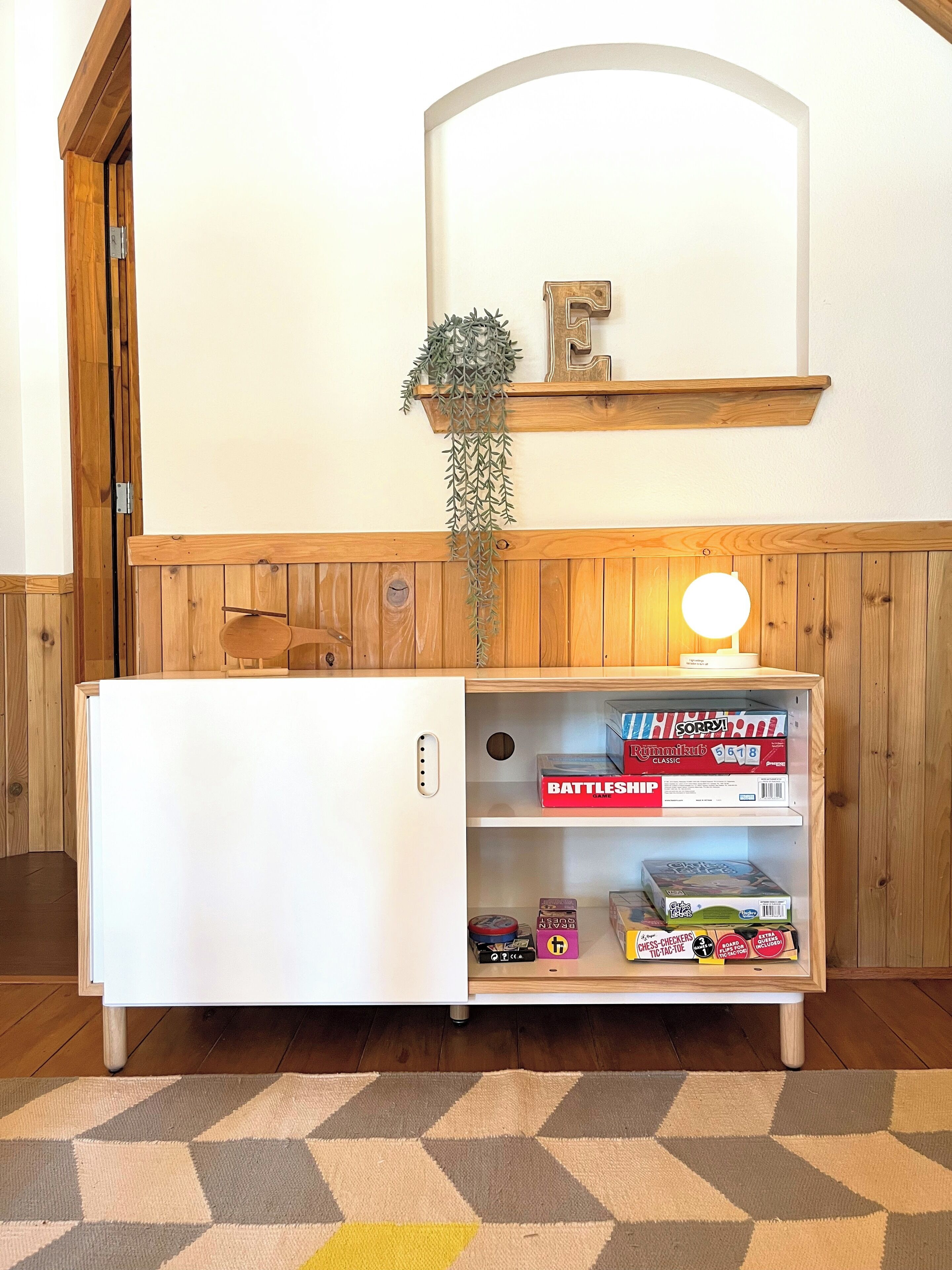 A compact room with a white refrigerator, a wooden bench, a shelf with board games, and a hanging plant.