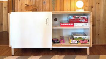 A compact room with a white refrigerator, a wooden bench, a shelf with board games, and a hanging plant.