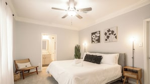 Een slaapkamer met een bed, een plafondventilator, een nachtkastje, een stoel, een plant en kunst aan de muur.