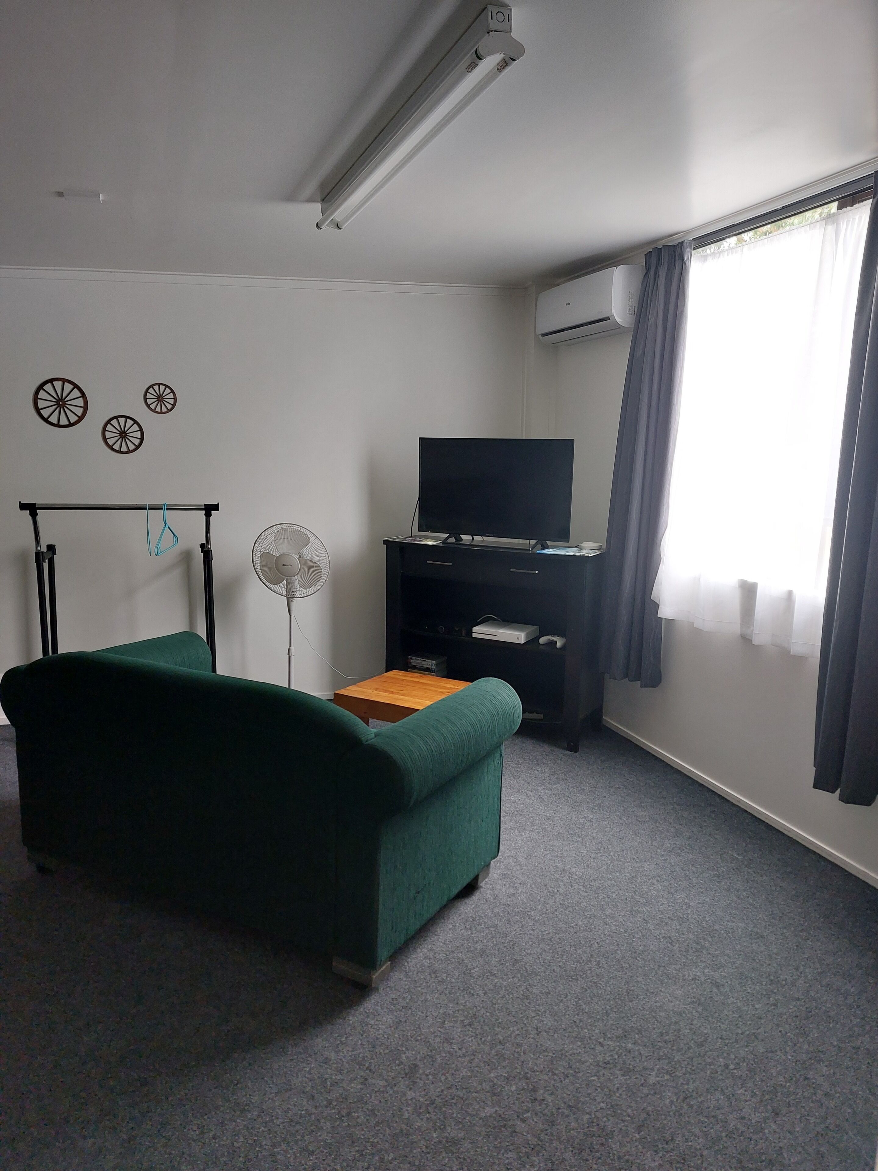 A room with a green sofa, a flat-screen TV on a stand, a small wooden table, a standing fan, and a window with curtains.