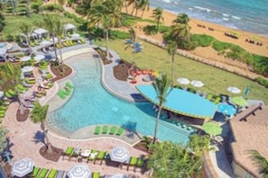 Ein Luftbild zeigt einen Resort mit großem Pool, Liegestühlen, Sonnenschirmen und einem Strandabschnitt.