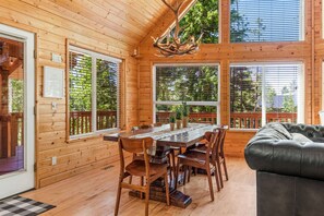 A wooden cabin interior with a dining table, chairs, a sofa, and large windows with blinds.