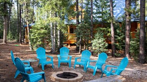 A forested area with a fire pit and six blue Adirondack chairs arranged around it.