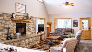 A cozy living room with a stone fireplace, comfortable seating, a television, and a large window.