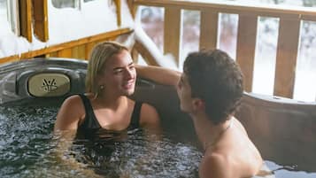 Zwei Personen in einer Whirlwanne, mit einer Holzterrasse und verschneiter Landschaft, die durch die Fenster sichtbar ist.