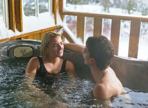 Dos personas en un jacuzzi con una terraza de madera y un paisaje nevado visible a través de las ventanas.