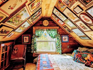 A cozy attic bedroom with a sloped ceiling adorned with framed artwork, a large window with curtains, a bed with a colorful quilt, and a chair with a decorative cushion. - Miracles on Polly - Memory Lane Cabin (Belfast)
