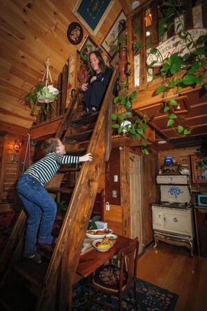 A wooden staircase with a person climbing it. There is a dining table with a plate of food and a chair. - Miracles on Polly - Memory Lane Cabin (Belfast)