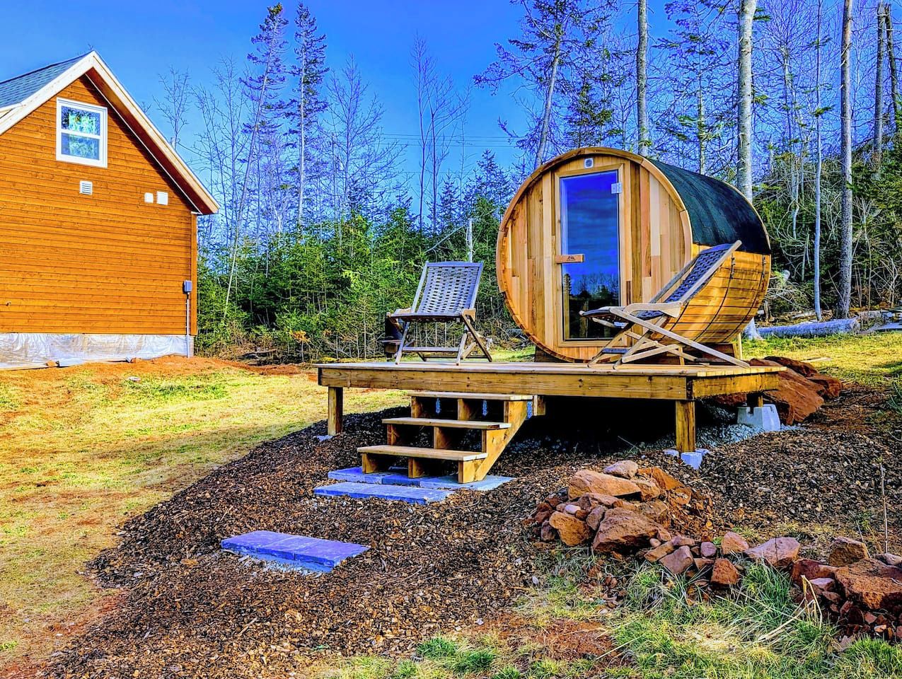 A wooden cabin with a sauna, a deck with chairs, and a small house in the background.