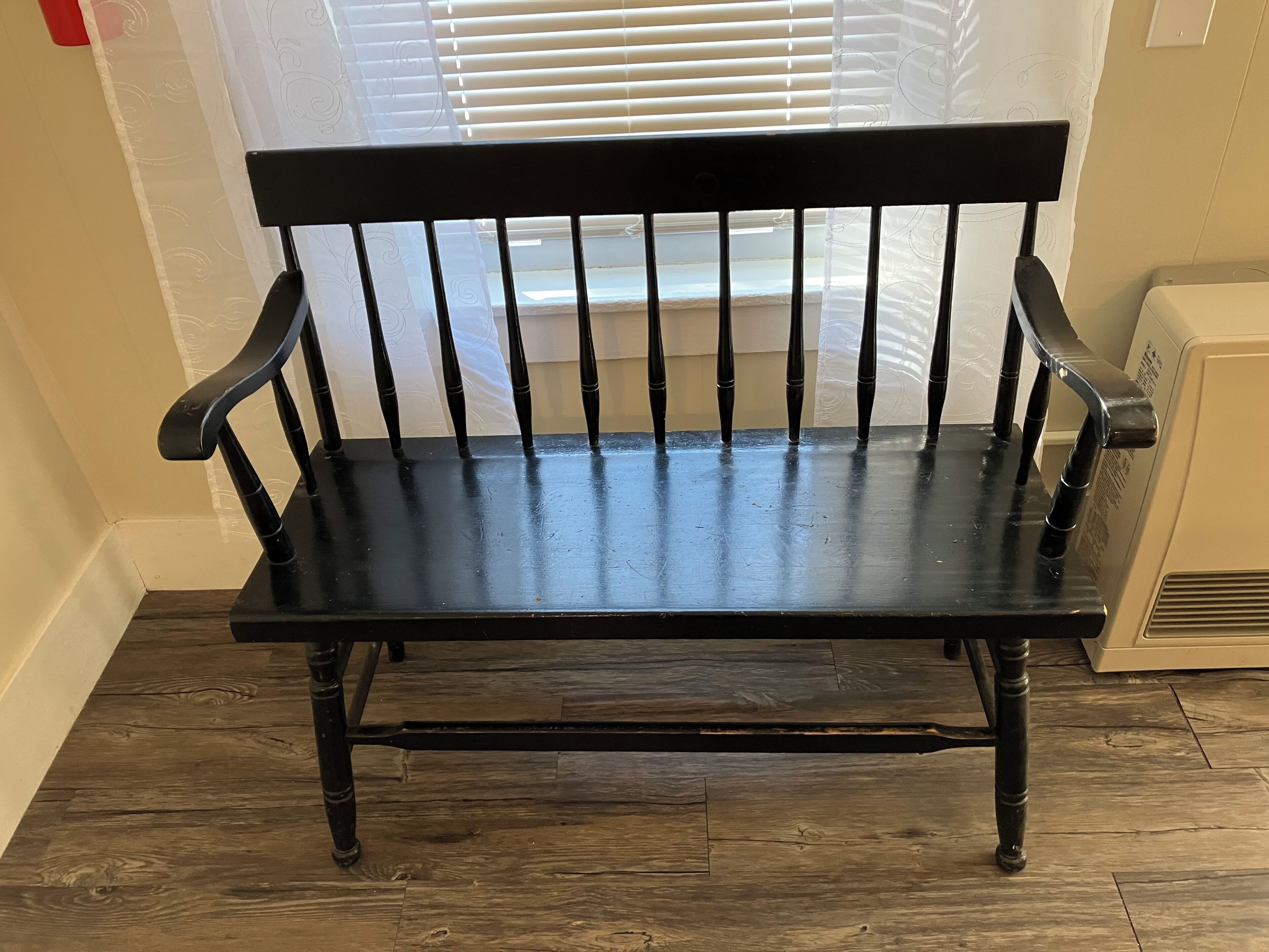 A black wooden bench with a backrest and armrests, positioned in a corner with a window in the background.