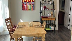 Un coin repas comprenant une table en bois, des chaises, un lustre, un tableau coloré et une étagère en métal où sont rangés des ustensiles de cuisine.