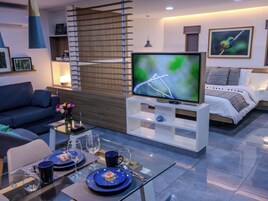 Un salón moderno con televisor de pantalla plana, un sofá, una mesa de comedor para dos personas y una cama con vistas al exterior.