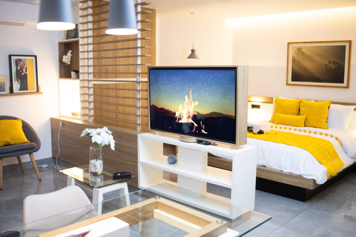 A modern hotel room with a bed, a TV, a glass coffee table, and a seating area with a chair and a small table.