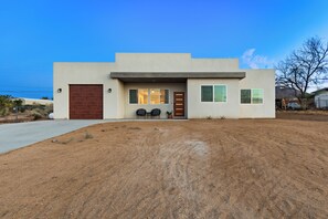 Uma casa moderna de dois andares com garagem, varanda frontal com cadeiras e um pequeno jardim.