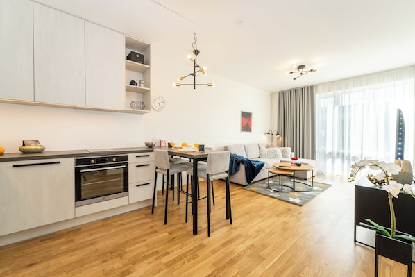 A modern kitchen with white cabinets, a built-in oven, and a dining area with a table and chairs. There is a living area with a sofa and a coffee table.