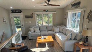 A spacious living room with a sectional sofa, a coffee table, and a fireplace. There is a ceiling fan, a large window, and a door labeled "BEACH".