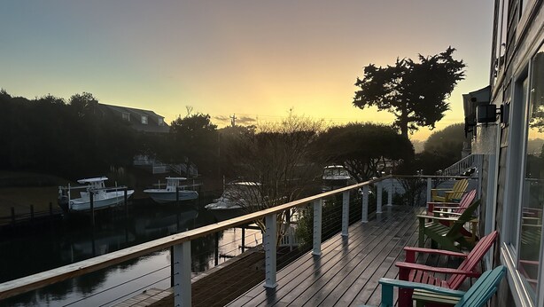 A wooden deck with colorful Adirondack chairs, a view of boats on a calm water body, and a clear sky.