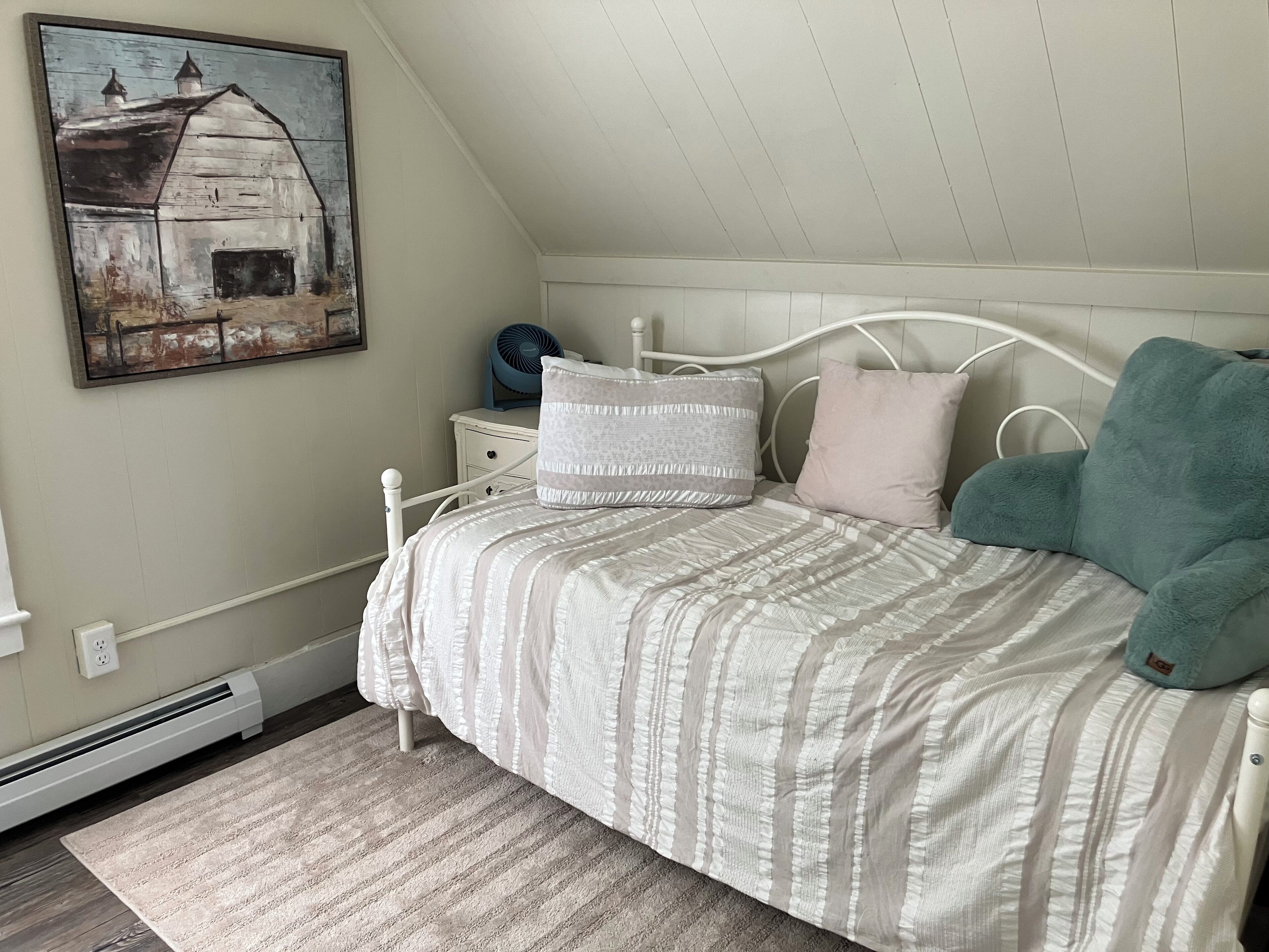 Ein Schlafzimmer mit Bett, Kissen, Nachttisch, Ventilator und einem Bild an der Wand.