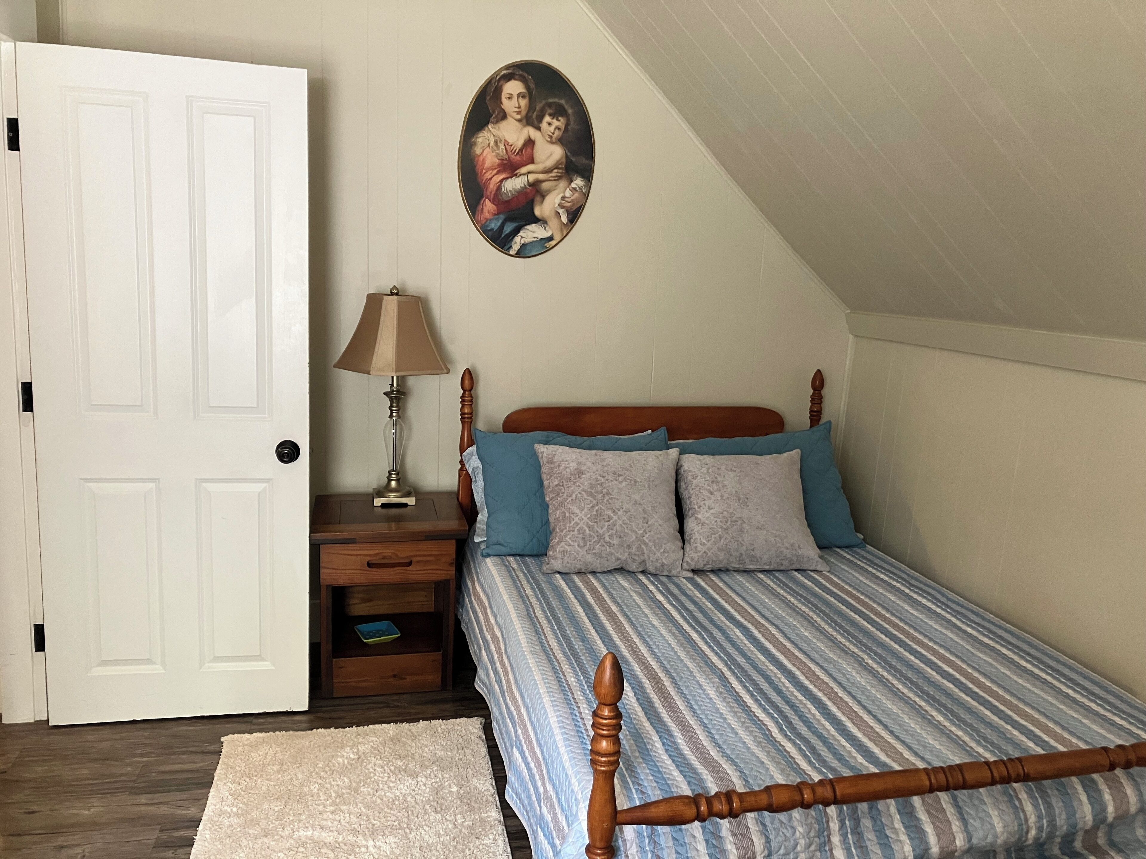 Een slaapkamer met een houten bed, een nachtkastje, een lamp, een ingelijst schilderij aan de muur en een witte deur.