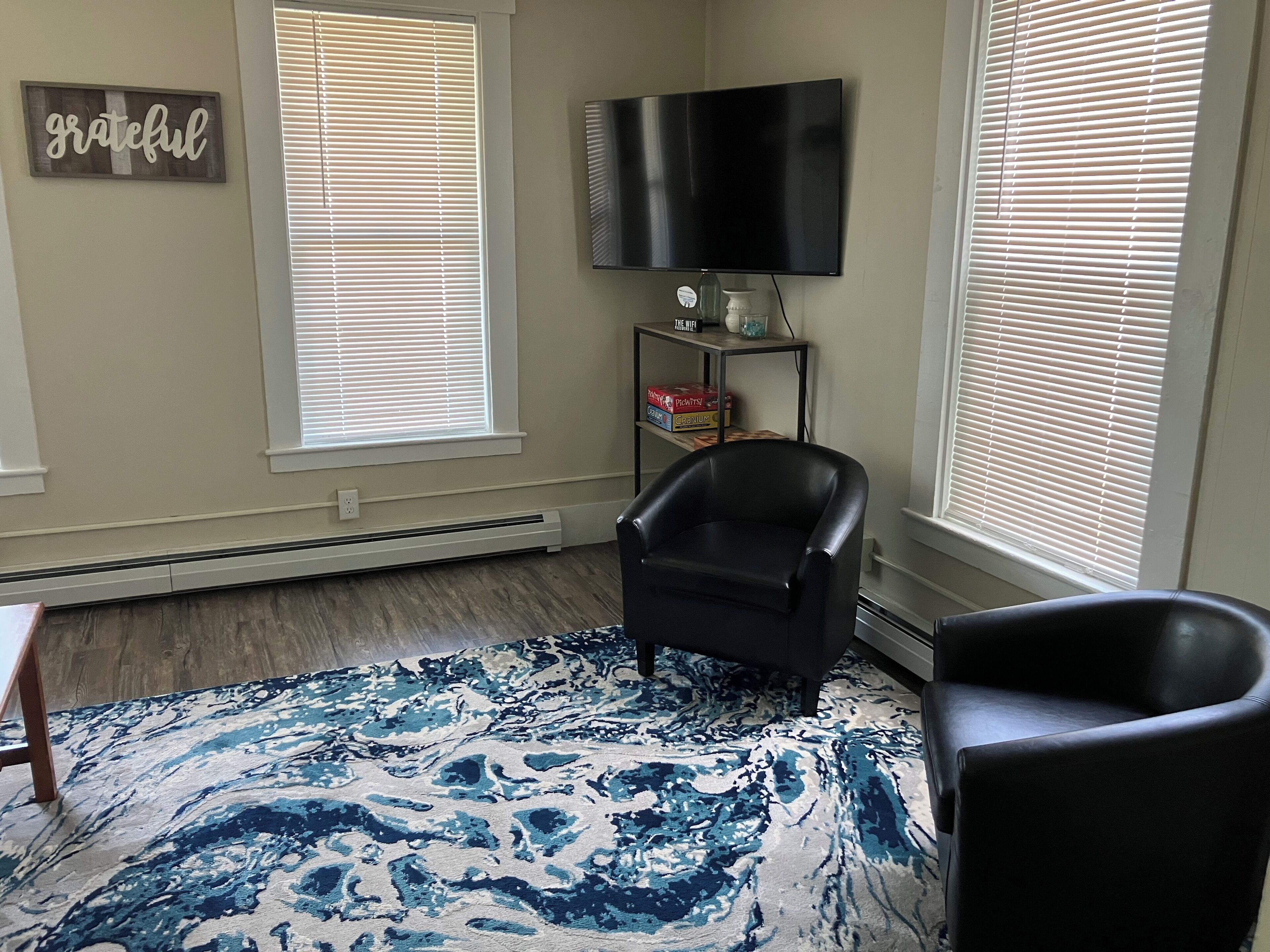 A living room with a black leather chair, a blue patterned rug, a flat-screen TV, and a 'grateful' sign on the wall.