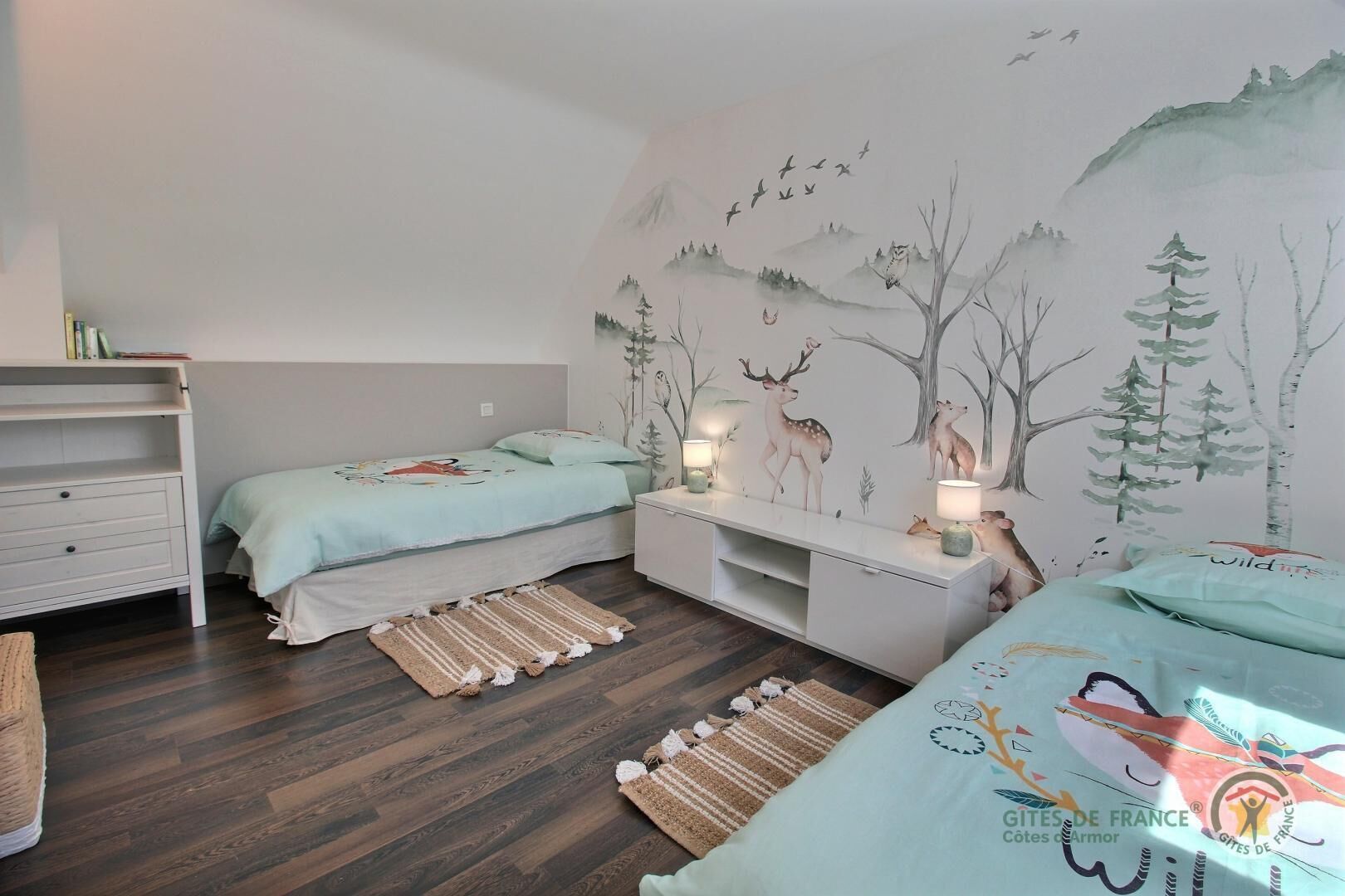 A bedroom with two beds, a nightstand, a dresser, and a mural of forest animals.