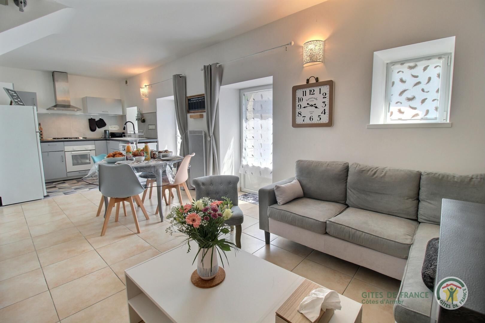 A modern living room with a grey sofa, a dining area with a table and chairs, a kitchen with white cabinets, and a clock on the wall.