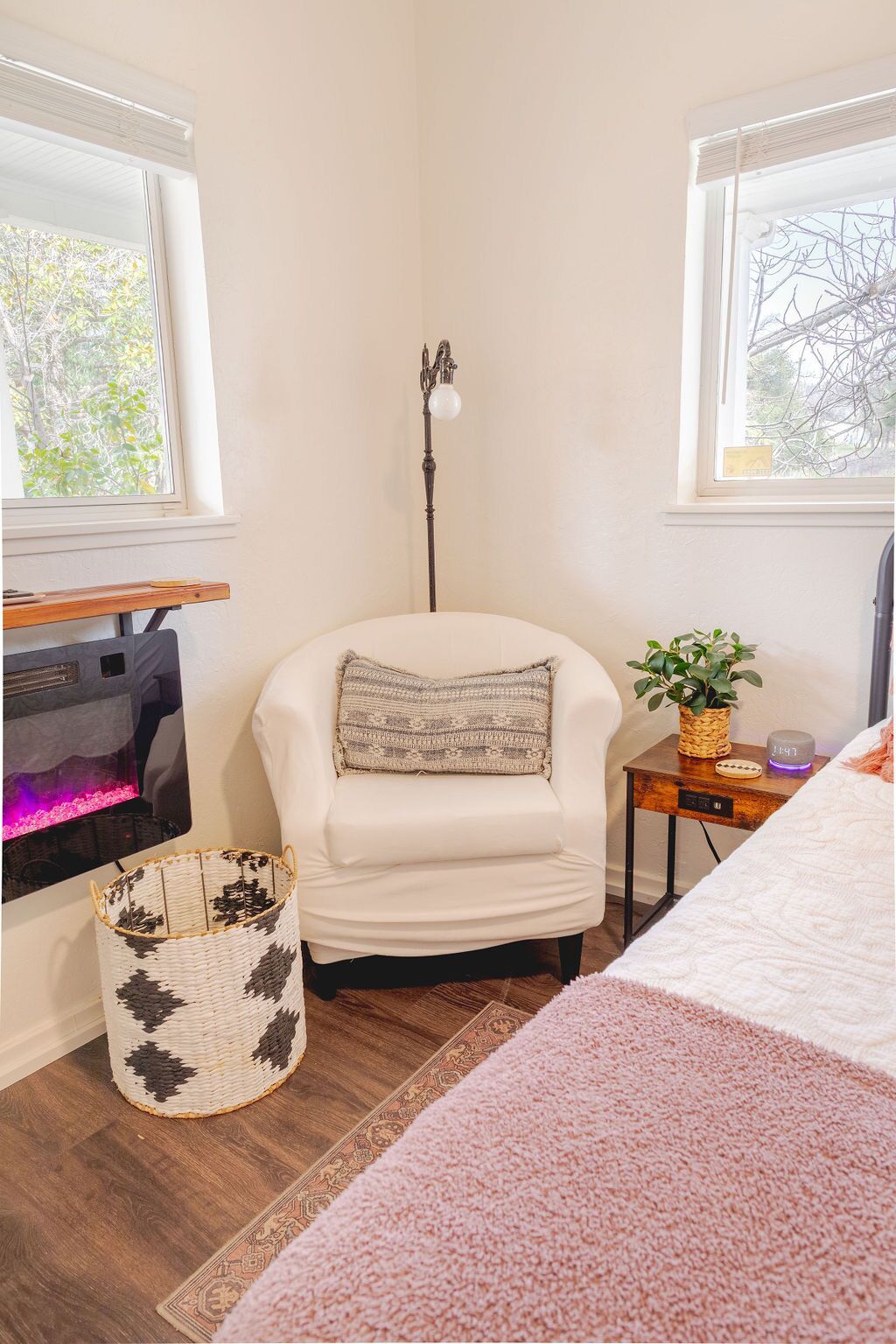 A cozy room with a white armchair, a small side table with a plant, a woven basket, and a pink rug.