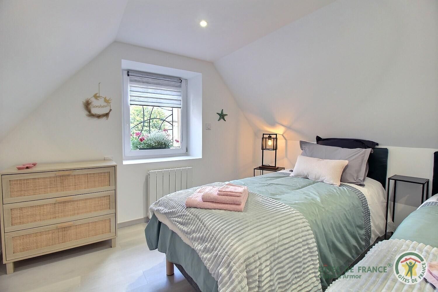 Quarto com teto inclinado, janela com vista para vegetação, mesa de cabeceira com abajur, cômoda e cama com manta listrada.