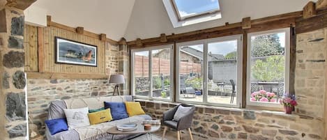Ein gemütliches Zimmer mit Steinwänden, einer Holzbalkendecke und großen Fenstern mit Blick auf den Garten.