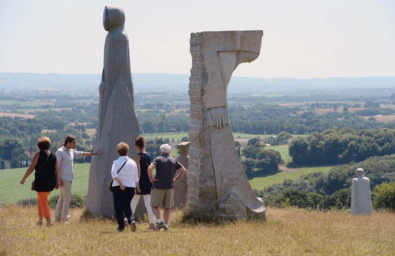 Eine Gruppe von Menschen betrachtet ein großes, markantes Steinbauwerk vor einer ländlichen Kulisse mit Feldern und Bäumen.