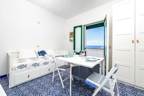 Ein kleiner, heller Raum mit einem Bett, einem Tisch und einem Stuhl. Es gibt ein Fenster mit Meerblick.
