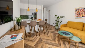 Una sala moderna con piso de madera en diseño de espiga, un sofá amarillo y una mesa de centro de vidrio. Incluye una zona de cocina con armarios blancos y lámparas colgantes amarillas.