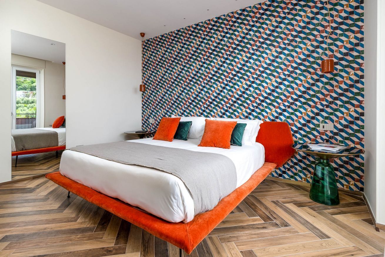 Une chambre moderne avec un mur orné d’un motif géométrique, un grand lit avec des coussins orange et verts, et un parquet en bois disposé en chevrons.
