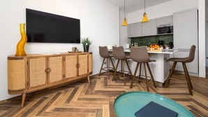 Una cocina moderna con armarios blancos, un comedor con cuatro sillas, un televisor de pantalla plana grande y un suelo de madera con diseño de espiga.