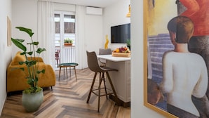 Una sala de estar moderna con un sofá, una zona de comedor con mesa y sillas, una cocina con televisor, y un cuadro en la pared.