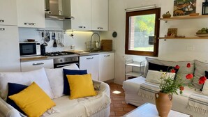 Een knus woongedeelte met een kitchenette, een bank met gele kussens, een kleine tafel met een vaas, en een raam met uitzicht op groen.