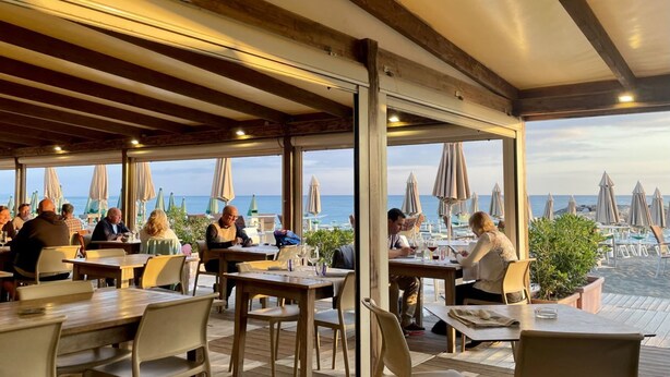 Een buitenruimte met eettafels en stoelen, grote parasols en uitzicht op zee.