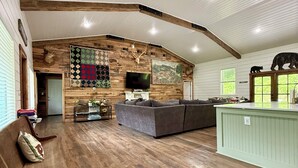 A spacious living room with wooden floors, a large gray sofa, a wooden bench, and a green island counter. - 55 ACRE WATERFRONT RETREAT ~HOT TUB~SWIM ~FISH ~KAYAK ~HIKE ~1 ACRE SPRING POND~ (Columbia)