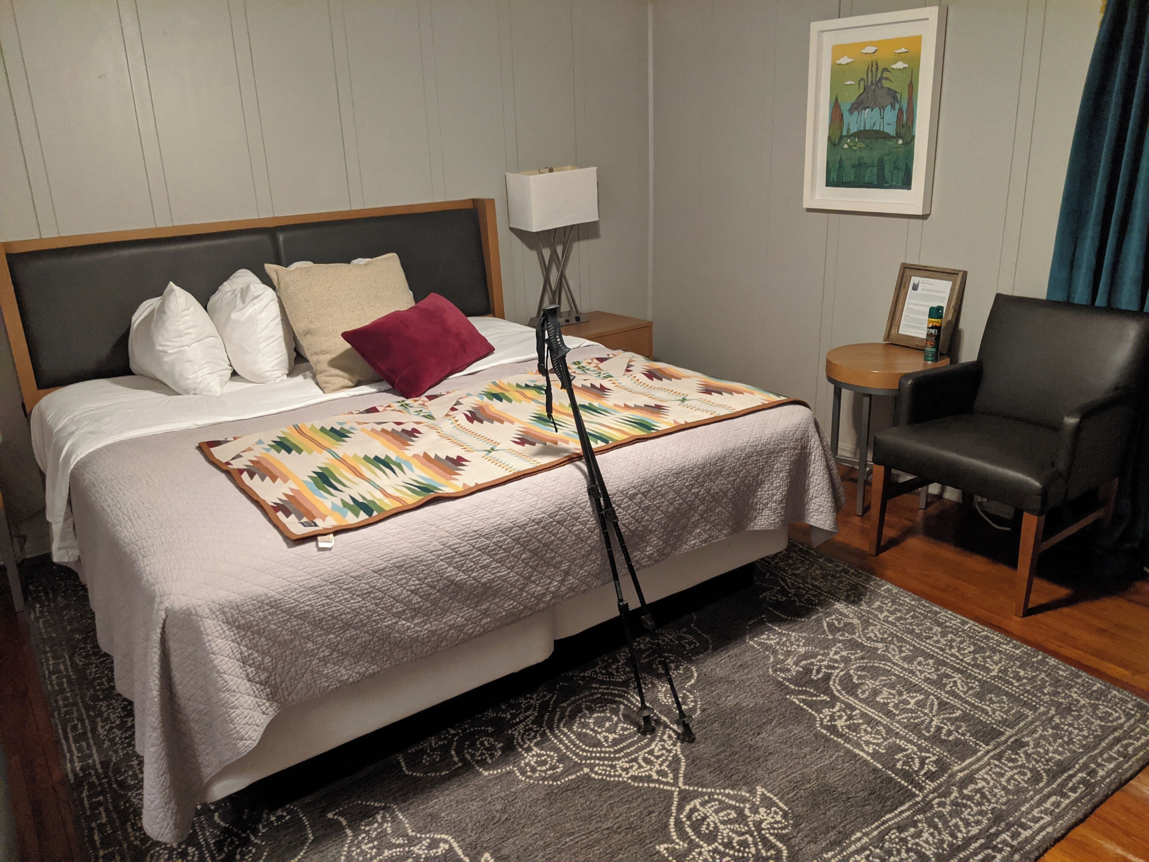 A neatly made bed with a patterned blanket, a bedside lamp, a chair, and a small table with a bottle.