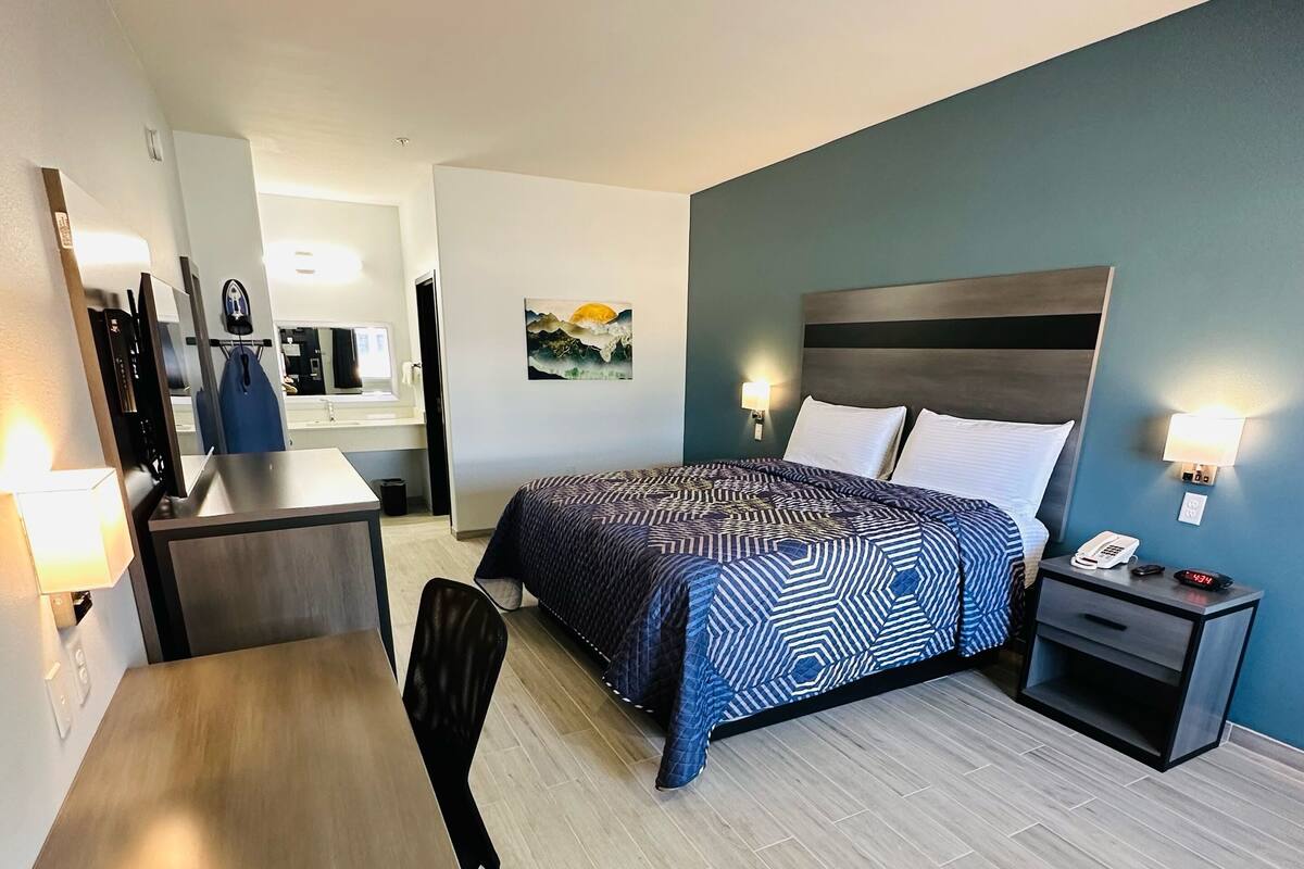 A hotel room with a bed, a desk, a chair, a nightstand, and a wall-mounted lamp.